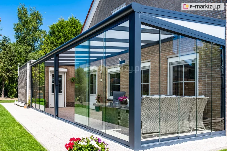 Narożne ujęcie całkowicie zabudowanego tarasu, opartego na konstrukcji aluminiowej w kolorze antracytowym, przylegającego do ściany budynku z czerwonej cegły. Zadaszenie posiada dach wykonany z mlecznego poliwęglanu, zamontowany na systemowych krokwiach, a boki oraz front konstrukcji zostały wypełnione przesuwnymi panelami ze szkła hartowanego, tworząc zamknięty ogród zimowy. Wewnątrz przeszklonej przestrzeni widoczna jest strefa wypoczynkowa z jasnoszarą, plecioną sofą narożną oraz stolikiem kawowym. Konstrukcja wspiera się na solidnych profilach pionowych, a całość posadowiona jest na podłożu z jasnego kruszywa, które graniczy z pasem białych kamieni ozdobnych i zadbanym trawnikiem. Wzdłuż bocznej ściany budynku przebiega szeroka ścieżka z jasnych płyt chodnikowych, prowadząca w głąb ogrodu z wysoką, zieloną roślinnością. Na elewacji domu widoczne są białe ramy okienne i drzwiowe z zewnętrznymi roletami, a na pierwszym planie znajduje się donica z kolorowymi kwiatami.