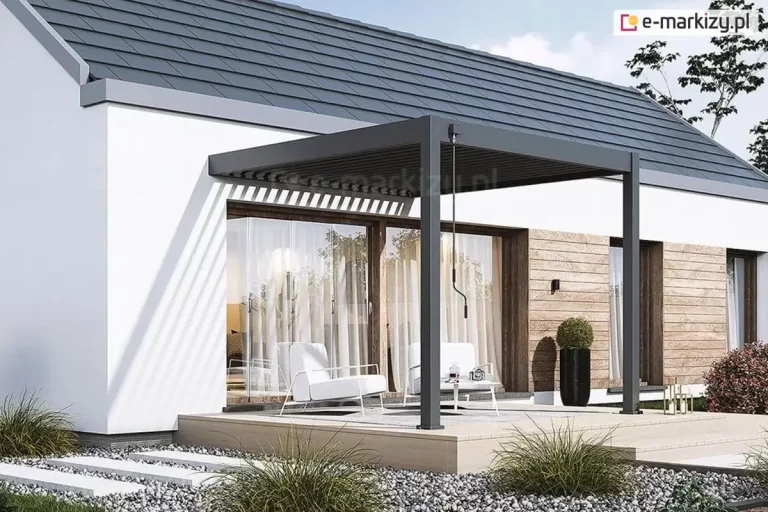 Antracytowa pergola lamelowa Marlo w wersji przyściennej, zamontowana na tarasie nowoczesnego domu parterowego z białym tynkiem i elementami drewnianej elewacji. Konstrukcja wspiera się na dwóch pionowych słupach o kwadratowym przekroju, a lamele dachowe są częściowo otwarte, co powoduje rzucanie pasowych cieni na ścianę budynku oraz przeszklone drzwi tarasowe. Przy lewym słupie widoczna jest czarna korba do ręcznej regulacji zadaszenia. Taras jest lekko podwyższony względem gruntu i wyłożony jasnymi płytami, a pod pergolą ustawiono dwa białe, nowoczesne fotele oraz niewielki stolik. Wokół tarasu znajduje się opaska z jasnego żwiru, w której osadzono betonowe płyty chodnikowe prowadzące do ogrodu, a budynek zwieńczony jest dwuspadowym dachem z ciemnoszarej dachówki.