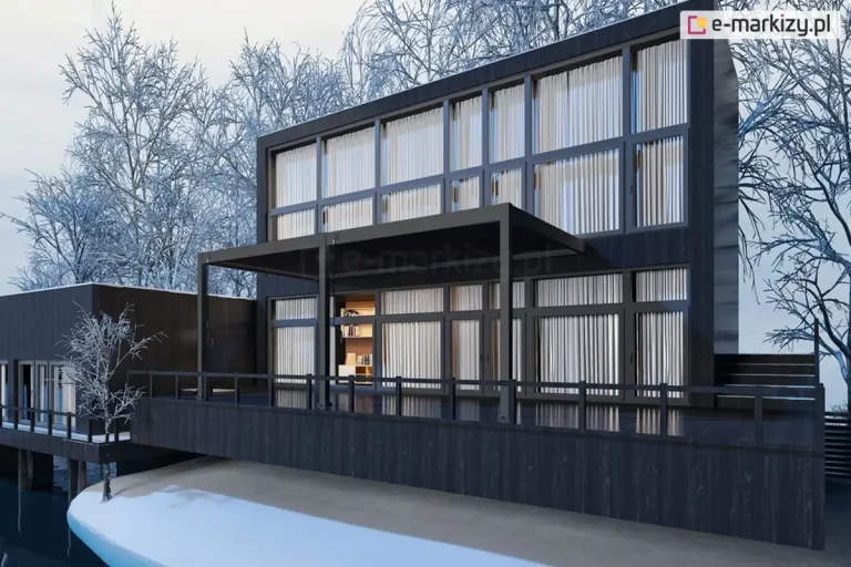 Dwumodułowa pergola bioklimatyczna Marlo w kolorze antracytowym, zainstalowana na rozległym tarasie nowoczesnego budynku o ciemnej, drewnianej elewacji. Konstrukcja zadaszenia jest zamontowana do ściany budynku i wspiera się na trzech pionowych słupach o kwadratowym przekroju. Lamele dachowe obu modułów są całkowicie zamknięte, tworząc płaską i szczelną powierzchnię. Budynek posiada liczne, wysokie przeszklenia z widocznymi jasnymi zasłonami wewnątrz. Taras jest wyniesiony ponad poziom gruntu, wykończony ciemnymi deskami i zabezpieczony minimalistyczną balustradą. Sceneria ma charakter zimowy – w tle widoczne są oszronione, bezlistne drzewa oraz zalegający na dole śnieg. Całość ujęcia prezentuje bryłę budynku z perspektywy bocznej, podkreślając integrację pergoli z nowoczesną architekturą domu.