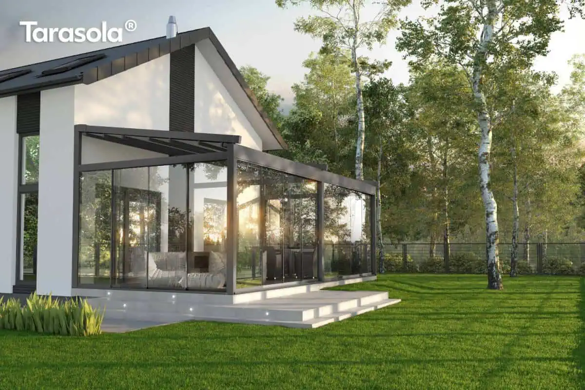 Nowoczesna, całoroczna zabudowa tarasu w formie przeszklonej pergoli Elegancy Tarasola o antracytowej konstrukcji aluminiowej, przytwierdzona do białej elewacji domu jednorodzinnego z dwuspadowym dachem. System wyposażony jest w pełne przeszklenia boczne z przesuwnymi panelami szklanymi, które tworzą zamknięty ogród letni. Wewnątrz przestrzeni rekreacyjnej znajdują się jasnoszare, miękkie pufy oraz strefa jadalniana z krzesłami w tle. Podstawa tarasu została wykonana z gładkich, jasnych płyt betonowych, w których stopniach zamontowano punktowe, dekoracyjne oświetlenie LED. Budynek posiada nowoczesne detale, takie jak ciemne pionowe pasy na elewacji i okna połaciowe. Całość otacza rozległy, zielony trawnik oraz lasek brzozowy, a w taflach szkła widoczne są refleksy zachodzącego słońca, podkreślające transparentność i estetykę konstrukcji.