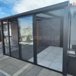 Nowoczesna konstrukcja ogrodu zimowego z pełnym przeszkleniem systemem ramowym Sotro. Grafitowe profile kontrastują z jasnymi płytami tarasowymi i szarą ścianą o strukturze kamienia wewnątrz zabudowy. Panele szklane są częściowo rozsunięte, co prezentuje łatwość obsługi wielotorowego systemu jezdnego.