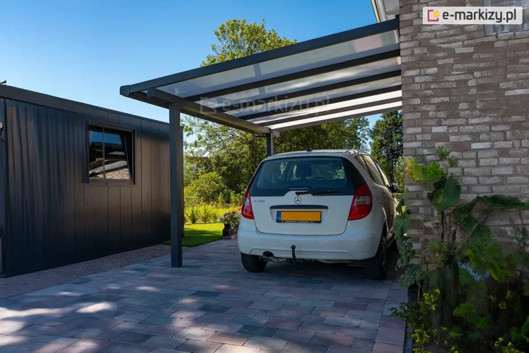 Zdjęcie przedstawia nowoczesną wiatę samochodową typu carport model Nebbiolo marki Deponti, zamontowaną przy bocznej ścianie budynku wykonanego z jasnej, klinkierowej cegły. Konstrukcja zadaszenia opiera się na ciemnoszarym, aluminiowym stelażu o matowym wykończeniu, składającym się z grubych profili nośnych oraz pionowego słupa zakotwiczonego w podłożu. Dach wiaty jest jednospadowy, nachylony pod niewielkim kątem w stronę zewnętrzną, a jego wypełnienie stanowią mleczne płyty poliwęglanowe o komorowej strukturze, które przepuszczają rozproszone światło dzienne. Pod zadaszeniem zaparkowany jest biały samochód osobowy marki Mercedes klasy A. Nawierzchnia pod wiatą oraz podjazd zostały wybrukowane betonową kostką w odcieniach szarości i brązu. W tle, po lewej stronie, widoczny jest fragment nowoczesnego budynku gospodarczego z elewacją z ciemnych paneli oraz przydomowy ogród z zieloną roślinnością i drzewami pod bezchmurnym, niebieskim niebem.