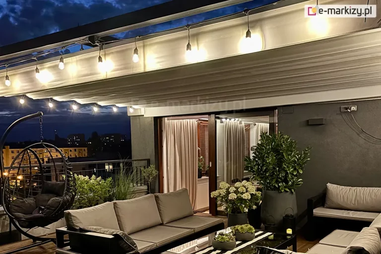 Zdjęcie przedstawia nocną aranżację luksusowego tarasu dachowego, nad którym zainstalowano system poziomego dachu rzymskiego Supro w kolorze jasnokremowym. Tkanina zadaszenia jest w pełni rozłożona i oświetlona od spodu girlandą świetlną z żarówkami o ciepłej barwie, co podkreśla regularne pofałdowania materiału. W centralnej części tarasu znajduje się strefa wypoczynkowa z nowoczesną, ciemną sofą wyposażoną w jasnoszare poduszki oraz niskim stolikiem kawowym z czarnym blatem. Po lewej stronie widoczny jest ażurowy fotel wiszący typu kokon, zawieszony na metalowym stelażu. Wnętrze apartamentu, widoczne przez otwarte przesuwne drzwi tarasowe, jest oświetlone i osłonięte jasnymi zasłonami. Na ścianie zewnętrznej o ciemnym tynku zamontowano minimalistyczny, czarny kinkiet świecący w dwóch kierunkach. Całość aranżacji uzupełniają liczne rośliny w donicach, w tym kwitnące na biało hortensje oraz ozdobne trawy. W tle, za metalową balustradą, widoczna jest panorama oświetlonego miasta pod nocnym, ciemnym niebem.