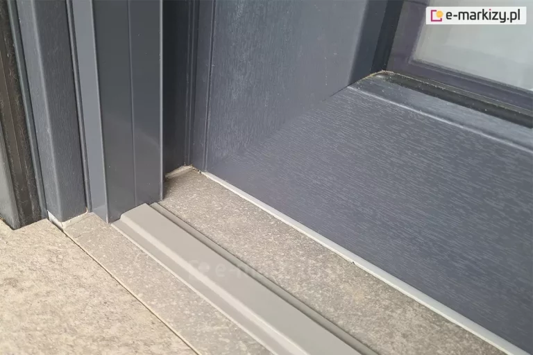 Próg dolny płaski moskitiery Click Rol zamocowany na płytkach gresowych w pobliżu drzwi balkonowych. Obok ramy okna oraz profilu moskitiery widoczne prowadnice rolety zewnętrznej z uszczelkami szczotkowymi.