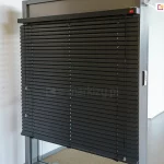 Widok na ekspozycyjny model żaluzji fasadowej C50 Slim w kolorze antracytowym, zamontowanej w systemowej ramie aluminiowej. Zdjęcie prezentuje niemal całkowicie rozwinięty system, w którym szerokie lamele C50 są ustawione w pozycji lekko otwartej, ukazując precyzyjny układ drabinek sznurkowych. Górna rynna żaluzji jest estetycznie wykończona dopasowaną kolorystycznie kasetką, na której znajduje się subtelny, czerwony logotyp producenta. Po prawej stronie żaluzji widoczny system ręcznego sznurkowego sterowania żaluzją. Po prawej stronie profilu montażowego widoczny jest dyskretny przełącznik sterowania automatycznego, najprawdopodobniej przeznaczony do sterowania innym systemem. Ekspozycja znajduje się w jasnym, minimalistycznym wnętrzu z białymi ścianami, co pozwala na dokładną ekspozycję detali technicznych konstrukcji oraz wysokiej jakości powłoki lakierniczej elementów aluminiowych.