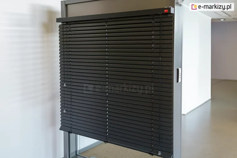 Widok na ekspozycyjny model żaluzji fasadowej C50 Slim w kolorze antracytowym, zamontowanej w systemowej ramie aluminiowej. Zdjęcie prezentuje niemal całkowicie rozwinięty system, w którym szerokie lamele C50 są ustawione w pozycji lekko otwartej, ukazując precyzyjny układ drabinek sznurkowych. Górna rynna żaluzji jest estetycznie wykończona dopasowaną kolorystycznie kasetką, na której znajduje się subtelny, czerwony logotyp producenta. Po prawej stronie żaluzji widoczny system ręcznego sznurkowego sterowania żaluzją. Po prawej stronie profilu montażowego widoczny jest dyskretny przełącznik sterowania automatycznego, najprawdopodobniej przeznaczony do sterowania innym systemem. Ekspozycja znajduje się w jasnym, minimalistycznym wnętrzu z białymi ścianami, co pozwala na dokładną ekspozycję detali technicznych konstrukcji oraz wysokiej jakości powłoki lakierniczej elementów aluminiowych.