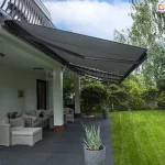 Perspektywa boczna na rozłożone osłony tarasowe - markizy Palladio Selt z poszyciem w kolorze ciemnoszarym, zamontowanymi pod sufitem balkonu nowoczesnego domu. Konstrukcja markiz opiera się na ramionach przegubowych, które utrzymują napięte, grafitowe poszycie nad strefą wypoczynku. Pod zadaszeniem widoczny jest komplet mebli ogrodowych z jasnej plecionki z miękkimi poduszkami, ustawiony na ciemnej, gładkiej posadzce tarasu. Białe filary budynku podtrzymują konstrukcję wyższej kondygnacji, a ściany wykończone są jasnym tynkiem. Obok mebli stoi wysoka, szara donica z ozdobną trawą, a w tle rozciąga się intensywnie zielony, równo przystrzyżony trawnik otoczony gęstą roślinnością ogrodową i wysokimi drzewami liściastymi. Scena oświetlona jest dziennym światłem, a na niebie widoczne są rozproszone, białe chmury.