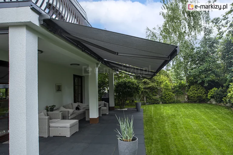 Perspektywa boczna na rozłożone osłony tarasowe - markizy Palladio Selt z poszyciem w kolorze ciemnoszarym, zamontowanymi pod sufitem balkonu nowoczesnego domu. Konstrukcja markiz opiera się na ramionach przegubowych, które utrzymują napięte, grafitowe poszycie nad strefą wypoczynku. Pod zadaszeniem widoczny jest komplet mebli ogrodowych z jasnej plecionki z miękkimi poduszkami, ustawiony na ciemnej, gładkiej posadzce tarasu. Białe filary budynku podtrzymują konstrukcję wyższej kondygnacji, a ściany wykończone są jasnym tynkiem. Obok mebli stoi wysoka, szara donica z ozdobną trawą, a w tle rozciąga się intensywnie zielony, równo przystrzyżony trawnik otoczony gęstą roślinnością ogrodową i wysokimi drzewami liściastymi. Scena oświetlona jest dziennym światłem, a na niebie widoczne są rozproszone, białe chmury.