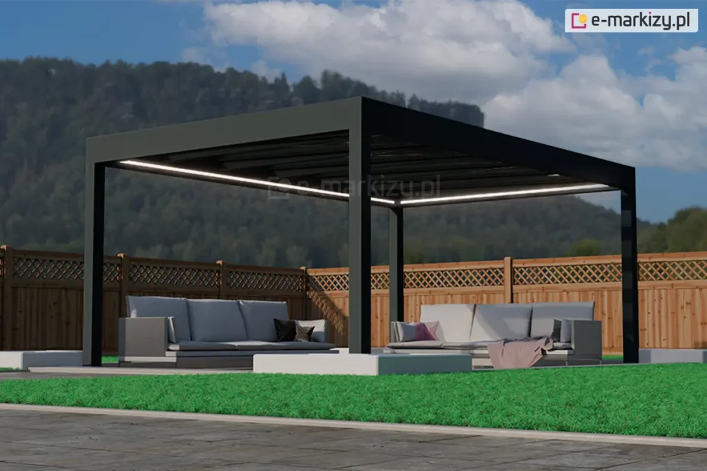 Wolnostojąca altana ogrodowa Spekta Glass o geometrycznej konstrukcji wykonanej z profili aluminiowych w kolorze głębokiej czerni. Rama nośna opiera się na czterech masywnych słupach o przekroju kwadratowym, a jej płaski dach posiada wypełnienie z dużych tafli szkła, które zapewniają ochronę przed opadami bez ograniczania dopływu światła. Wzdłuż wewnętrznych krawędzi konstrukcji zainstalowano system liniowego oświetlenia LED, emitujący ciągły pas intensywnego, białego światła. Pod zadaszeniem zorganizowano strefę wypoczynkową z dwiema dużymi sofami ogrodowymi w kolorze jasnoszarym, wyposażonymi w miękkie poduchy oraz dekoracyjny, różowy koc. Podłoże pod altaną stanowią gładkie, jasnoszare płyty tarasowe, które kontrastują z otaczającym je żywozielonym, krótko przystrzyżonym trawnikiem. W bezpośrednim sąsiedztwie słupów wkomponowano niskie, białe donice o geometrycznym kształcie, wypełnione drobnym, jasnym kruszywem. Tło kompozycji stanowi wysokie, drewniane ogrodzenie panelowe z ozdobnym, ażurowym wykończeniem w górnej części, za którym rozciąga się widok na zalesione wzgórze pod błękitnym niebem. Całość realizacji charakteryzuje się nowoczesnym designem i wysoką estetyką, tworząc luksusowe miejsce do rekreacji.