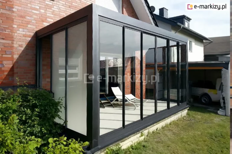 nowoczesny ogród zimowy o czarnej konstrukcji aluminiowej, dobudowany do ceglanej ściany budynku. Trzy ściany obiektu zostały przeszklone systemem Sotro, przy czym jedna z nich posiada mleczne wypełnienie zapewniające prywatność. Konstrukcja posadowiona jest na betonowym fundamencie, a we wnętrzu widoczne są leżaki ogrodowe. Minimalistyczny design czarnych profili kontrastuje z czerwienią cegły i zielenią trawnika otaczającego budowlę.