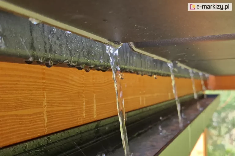 Techniczne zbliżenie na system odprowadzania wody opadowej w dachu lamelowym SB400 MD, zamontowanym w drewnianej konstrukcji pergoli. Na pierwszym planie widoczne są krawędzie aluminiowych lameli w ciemnym kolorze, z których spływają wyraźne strugi wody bezpośrednio do rynny bocznej. Rynna ma formę otwartego profilu o ciemnej barwie i jest zamontowana wzdłuż poziomej, masywnej belki wykonanej z drewna o wyraźnym usłojeniu i ciepłym odcieniu. Na dolnej krawędzi rynny oraz na bocznej płaszczyźnie drewnianej belki widoczne są liczne krople wody, co obrazuje działanie systemu w warunkach opadów deszczu. Tło jest lekko rozmyte, ukazując naturalną zieleń ogrodu, co pozwala skupić uwagę na mechanicznym aspekcie odprowadzania wody z powierzchni zadaszenia.
