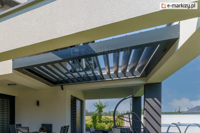 Zdjęcie prezentuje detal montażowy dachu lamelowego Selt SB400R w wersji z ramą, zainstalowanego w przygotowanym otworze żelbetowego zadaszenia tarasu. Aluminiowa konstrukcja w kolorze antracytowym jest ściśle dopasowana do białej wnęki stropu, tworząc estetyczną i zlicowaną powierzchnię. Ruchome pióra pergoli są otwarte, co ukazuje mechanizm obrotowy oraz pozwala na swobodny przepływ światła i wentylację przestrzeni pod spodem. Na pierwszym planie widoczny jest narożnik konstrukcji dachu z precyzyjnymi łączeniami oraz fragment rynny odprowadzającej wodę. Poniżej zadaszenia znajduje się strefa wypoczynkowa z ciemnymi meblami ogrodowymi, żaluzjami fasadowymi na oknach budynku oraz wiszącym fotelem. W tle widać otwartą przestrzeń ogrodu z zielenią i błękitne niebo. Na ścianie budynku zamontowano minimalistyczne oświetlenie zewnętrzne, a jasna elewacja kontrastuje z ciemnymi elementami ślusarki aluminiowej.