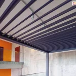 Widok od dołu na zadaszenie lamelowe pergoli SB350 Aluprof w kolorze antracytowym, zamontowanej wewnątrz nowoczesnego budynku. Ruchome lamele są ustawione pod kątem, ukazując biały sufit z zamontowaną szyną oświetleniową i białymi reflektorami punktowymi. Konstrukcja pergoli opiera się na ciemnych słupach i przylega do betonowej ściany o surowej teksturze. W tle widoczna jest antresola z szklaną balustradą, fragment intensywnie pomarańczowej ściany oraz drzwi w kolorze ciemnego drewna. Perspektywa żabia podkreśla techniczne detale mechanizmu obrotowego lameli oraz integrację konstrukcji z architekturą wnętrza.