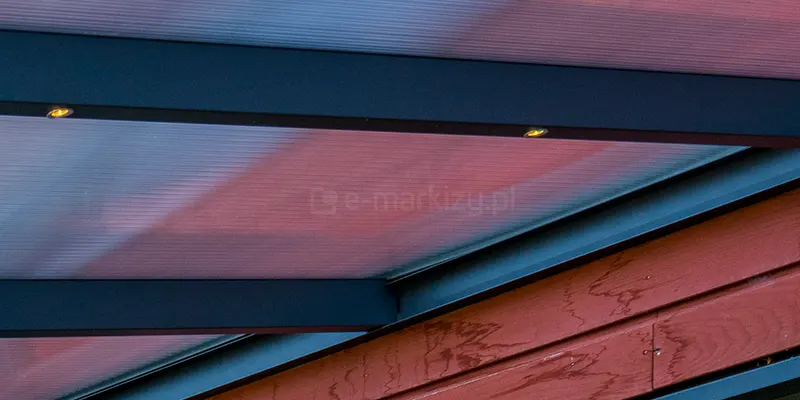 Konstrukcja aluminiowa mocowana do ściany z widocznymi w krokwi oczkami punktowego oświetlenia LED. W antracytowej konstrukcji zamocowane płyty poliwęglanu komorowego 16mm w barwie przejrzystej, przez którą przebija czerwonawy kolor drewnianej ściany.