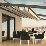 Nowoczesny dach rzymski Square zamontowany na przestronnym tarasie apartamentu. Konstrukcja zadaszenia opiera się na solidnych, aluminiowych profilach w kolorze antracytowym, które są przytwierdzone do elewacji budynku i wsparte na poprzecznych belkach. Jasnobeżowa, wodoodporna tkanina jest częściowo rozwinięta, tworząc charakterystyczne, głębokie fałdy podwieszone pod prowadnicami. Pod zadaszeniem znajduje się strefa jadalniana z dużym białym stołem oraz kompletem ośmiu nowoczesnych krzeseł w kolorze czarnym z białymi nogami. Taras wykończony jest jasnymi deskami kompozytowymi o wysokim połysku, w których odbijają się meble i elementy konstrukcyjne. W tle rozpościera się szeroka panorama miasta widoczna z dużej wysokości, ograniczona minimalistyczną balustradą ze stalowych linek. Na ścianie budynku, obok dużych przeszkleń, zamontowane są klasyczne kinkiety zewnętrzne, a całość oświetlona jest ciepłym światłem zachodzącego słońca, co podkreśla ekskluzywny charakter aranżacji.