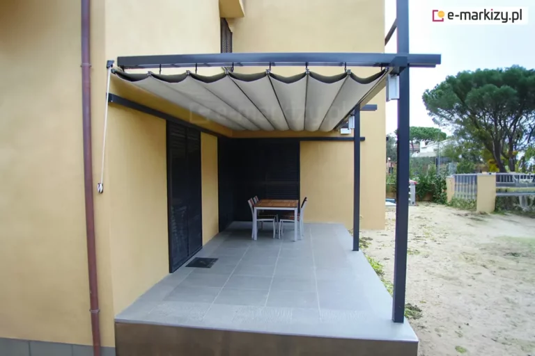 system zadaszenia rzymskiego Silva ze sterowaniem ręcznym, zamontowany na tarasie przylegającym do budynku o żółtej elewacji. Konstrukcja wsporcza zadaszenia składa się z ciemnoszarych, metalowych profili oraz dwóch pionowych słupów zamontowanych na krawędzi tarasu. Poszycie dachowe w kolorze beżowym jest w pełni rozłożone i podwieszone pod szynami, tworząc system regularnych, miękkich fal. Po lewej stronie, przy ścianie budynku, widoczny jest biały sznurek służący do ręcznej obsługi mechanizmu rozsuwania i zwijania tkaniny. Taras wyłożony jest dużymi, jasnoszarymi płytami gresowymi, a w jego centralnej części ustawiono prostokątny stół jadalniany z krzesłami. W tle widoczne są ciemne drzwi tarasowe oraz fragment niezagospodarowanego ogrodu z piaszczystym podłożem i drzewami iglastymi w oddali.