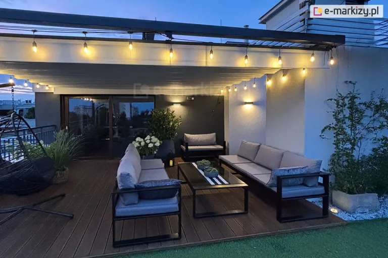 Dach rzymski Supro na prowadnicach zamocowany do aluminiowej konstrukcji okalającej taras na piętrze budynku. Dach otaczają girlandy światełek, nadając przestrzeni wyjątkowy klimat także po zmroku. Pod dachem, na drewnianym tarasie ustawiono zestaw mebli wypoczynkowych: dwie ławki z siedziskami oraz fotel i stolik, a także fotel typu kokon po lewej stronie. Pomiędzy tym umieszczono kilka donic z roślinami zielonymi i kwiatami.