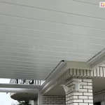 Zdjęcie prezentuje zaawansowane technicznie zadaszenie lamelowe SB400R firmy Selt w kolorze białym, zamontowane na tarasie o nieregularnym kształcie. Konstrukcja charakteryzuje się precyzyjnym docięciem w kształt litery L, co umożliwia idealne dopasowanie dachu do uskoku bryły budynku wykonanej z jasnej cegły klinkierowej. Aluminiowe lamele są ustawione w pozycji zamkniętej, tworząc jednolitą, szczelną i płaską powierzchnię sufitu, która skutecznie chroni przed opadami. Widoczne są rynny boczne zintegrowane z ramą zadaszenia, które odpowiadają za systemowy odpływ wody. Całość konstrukcji wspiera się na białych profilach aluminiowych, a na elewacji budynku, tuż pod linią dachu, zamontowane są zewnętrzne czujniki pogodowe lub punkty oświetleniowe. W tle widoczny jest fragment balkonu z metalową balustradą oraz zarys drzew, co podkreśla funkcjonalność systemu w zabudowie jednorodzinnej.