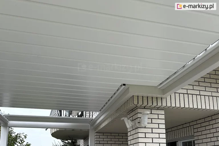 Zdjęcie prezentuje zaawansowane technicznie zadaszenie lamelowe SB400R firmy Selt w kolorze białym, zamontowane na tarasie o nieregularnym kształcie. Konstrukcja charakteryzuje się precyzyjnym docięciem w kształt litery L, co umożliwia idealne dopasowanie dachu do uskoku bryły budynku wykonanej z jasnej cegły klinkierowej. Aluminiowe lamele są ustawione w pozycji zamkniętej, tworząc jednolitą, szczelną i płaską powierzchnię sufitu, która skutecznie chroni przed opadami. Widoczne są rynny boczne zintegrowane z ramą zadaszenia, które odpowiadają za systemowy odpływ wody. Całość konstrukcji wspiera się na białych profilach aluminiowych, a na elewacji budynku, tuż pod linią dachu, zamontowane są zewnętrzne czujniki pogodowe lub punkty oświetleniowe. W tle widoczny jest fragment balkonu z metalową balustradą oraz zarys drzew, co podkreśla funkcjonalność systemu w zabudowie jednorodzinnej.