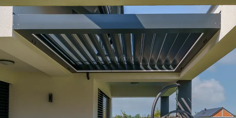 dach lamelowy Selt SB400R w kolorze antracytowym, zamontowany w przygotowanej wnęce betonowego zadaszenia tarasu. Aluminiowa rama pergoli jest precyzyjnie wbudowana w biały, otynkowany strop, tworząc estetyczne obramowanie dla ruchomych piór. Lamele znajdują się w pozycji otwartej, ustawione pod kątem, co ukazuje ich profil oraz pozwala na wgląd w błękitne niebo ponad konstrukcją. System jest wyposażony w zintegrowane oświetlenie LED w postaci listwy biegnącej wzdłuż wewnętrznej krawędzi ramy. Pod zadaszeniem widoczna jest jasna elewacja budynku z czarnymi detalami, takimi jak żaluzje fasadowe i nowoczesny kinkiet. W głębi tarasu znajduje się wiszący fotel ogrodowy oraz widok na otwartą przestrzeń z zielenią i sąsiednią zabudową.
