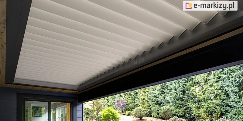 Zbliżenie na system dachu lamelowego Selt SB400R w konfiguracji z białymi lamelami i ciemnoantracytową ramą. Aluminiowe pióra dachu są ustawione w pozycji lekko uchylonej, co tworzy charakterystyczną, schodkową strukturę powierzchni i sugeruje możliwość precyzyjnej regulacji dopływu światła oraz cyrkulacji powietrza. Konstrukcja ramy jest zintegrowana z bryłą budynku, a pod nią widoczny jest fragment drewnianej podbitki sufitowej. W dolnej części kadru widać przeszklone drzwi tarasowe z widokiem na oświetlone wnętrze salonu oraz gęstą, zieloną roślinność ogrodu z licznymi drzewami iglastymi i krzewami. Perspektywa z dołu podkreśla rytmiczny układ lameli oraz nowoczesny design systemu zadaszenia.