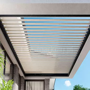 Bioklimatyczny dach lamelowy Bioline z otwartymi piórami wpuszczającymi światło, osadzony w minimalistycznej ramie betonowej na tarasie, za konstrukcją widać fragment elewacji domu oraz błękitne niebo tworzące lekką, wakacyjną atmosferę tarasu.