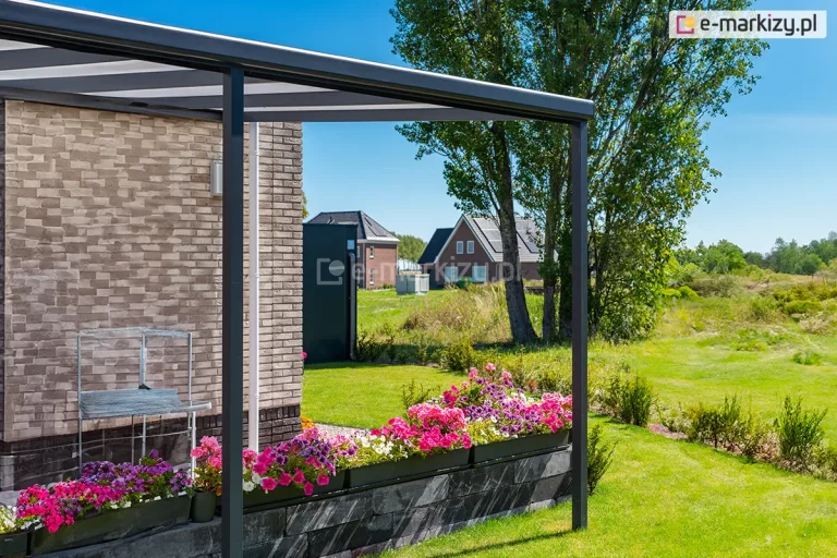 Zdjęcie prezentuje przyścienne zadaszenie tarasowe Nebbiolo marki Deponti, wykonane z wytrzymałych profili aluminiowych w kolorze antracytowym. Konstrukcja składa się z rynny frontowej, krokwi oraz smukłych słupów nośnych o kwadratowym przekroju, z których jeden jest widoczny na pierwszym planie, wsparty na podłożu przy krawędzi tarasu. Pokrycie dachu stanowią płyty z poliwęglanu komorowego o mlecznym wybarwieniu, które zapewniają ochronę przed opadami przy jednoczesnym przepuszczaniu zmiękczonego światła słonecznego. Zadaszenie przylega do elewacji budynku wyłożonej dekoracyjną płytką klinkierową w piaskowych i brązowych odcieniach. Taras wykończony jest murkiem z ciemnych bloków skalnych, na którym ustawiono podłużne donice z gęsto kwitnącymi kwiatami w kolorach różowym, fioletowym i białym. W tle rozpościera się widok na zadbany trawnik, wysokie drzewa liściaste oraz inne domy jednorodzinne o spadzistych dachach, widoczne pod czystym, błękitnym niebem.