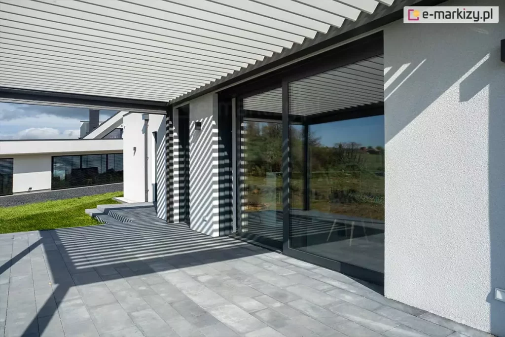 Widok z poziomu tarasu na przyścienną pergolę bioklimatyczną Sb400 Selt z białymi, aluminiowymi lamelami. Konstrukcja zadaszenia jest zintegrowana z nowoczesnym budynkiem o białej, tynkowanej elewacji i dużych przeszkleniach w ciemnoszarych ramach. Światło słoneczne przechodzące przez uchylone lamele rzuca wyraźny, geometryczny wzór pasów na ścianę domu oraz podłoże wykonane z dużych, szarych płyt tarasowych. W szybie przesuwnych drzwi tarasowych widoczne jest odbicie krajobrazu oraz dalszej części konstrukcji. W tle rozpościera się widok na zielony ogród, trawnik oraz sąsiedni budynek o minimalistycznej architekturze. Zdjęcie prezentuje efekt wizualny gry światła i cienia uzyskiwany dzięki regulacji kąta nachylenia lameli w pergoli.