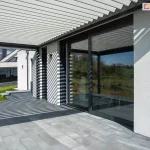 Widok z poziomu tarasu na przyścienną pergolę bioklimatyczną Sb400 Selt z białymi, aluminiowymi lamelami. Konstrukcja zadaszenia jest zintegrowana z nowoczesnym budynkiem o białej, tynkowanej elewacji i dużych przeszkleniach w ciemnoszarych ramach. Światło słoneczne przechodzące przez uchylone lamele rzuca wyraźny, geometryczny wzór pasów na ścianę domu oraz podłoże wykonane z dużych, szarych płyt tarasowych. W szybie przesuwnych drzwi tarasowych widoczne jest odbicie krajobrazu oraz dalszej części konstrukcji. W tle rozpościera się widok na zielony ogród, trawnik oraz sąsiedni budynek o minimalistycznej architekturze. Zdjęcie prezentuje efekt wizualny gry światła i cienia uzyskiwany dzięki regulacji kąta nachylenia lameli w pergoli.
