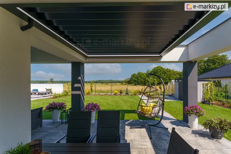 alty Niestandardowy Gem Fotografia prezentuje techniczne detale montażu dachu tarasowego SB400R, skupiając się na systemie odprowadzania wody opadowej oraz integracji z konstrukcją budynku. Na zbliżeniu widoczny jest czarny element odpływowy zamontowany do jasnej elewacji, który łączy wewnętrzny system rynnowy pergoli z pionem spustowym. Zadaszenie składa się z aluminiowych lameli w kolorze antracytowym, które tworzą szczelną płaszczyznę w pozycji zamkniętej. Wzdłuż wewnętrznej ramy konstrukcji zainstalowano liniowe oświetlenie LED w formie białych pasów, które równomiernie doświetlają przestrzeń wypoczynkową. Taras został wyłożony szarymi płytami kamiennymi, a na jego powierzchni znajdują się meble ogrodowe, wiszący fotel oraz ozdobne donice z kwiatami. Całość instalacji osadzona jest na masywnych, ciemnych filarach, które stanowią element konstrukcyjny bryły domu, otwartej na ogród z zielonym trawnikiem i niską roślinnością.