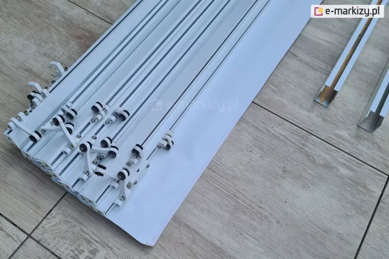 Uzbrojone belki przeciwwietrzne - zamocowane wózki - do dachu Supro, jako opcjonalnego zadaszenia pergoli drewnianej Moza. Belki z tkaniną ułożono w pobliżu prowadnic dachu na podłodze tarasowej wyłożonej płytkami gresowymi z drewnopodobnym wzorem.