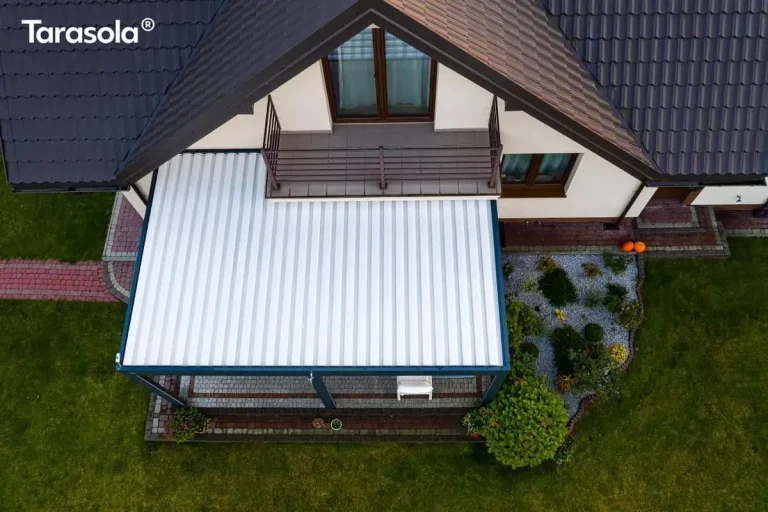 Zdjęcie wykonane z perspektywy lotu ptaka (widok prostopadły z góry) ukazuje kompletną połać dachową nowoczesnej pergoli Essential Tarasola zainstalowanej przy elewacji domu jednorodzinnego. Zadaszenie modelu Essential Tarasola składa się z szerokich, aluminiowych lameli w kolorze białym, które w tej pozycji są całkowicie zamknięte, tworząc jednolitą, żebrowaną płaszczyznę chroniącą taras przed opadami. Biały dach kontrastuje z ciemnoantracytową ramą konstrukcyjną oraz ciemnobrązową dachówką budynku. Pergola Essential Tarasola jest zamontowana bezpośrednio pod balkonem o brązowej balustradzie, przylegając do jasnej, beżowej ściany domu. Pod zadaszeniem widoczny jest fragment tarasu wyłożonego szarą kostką brukową oraz białe meble ogrodowe. Po prawej stronie konstrukcji znajduje się dekoracyjna rabata ogrodowa o nieregularnym kształcie, wysypana szarym gresem i obsadzona ozdobnymi krzewami, w tym dużą zieloną hortensją przy samym narożniku tarasu. Na styku opaski wokół domu a trawnikiem widoczne są dwie pomarańczowe dynie, sugerujące jesienną porę wykonania zdjęcia. Po lewej stronie kadru biegnie ścieżka z czerwonej kostki brukowej, otoczona zadbanym, zielonym trawnikiem.