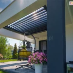 Widok na nowoczesny taras z zamontowanym w opasce betonowej wspartej na szarym masywnym słupie dachem lamelowym SB400R. Aluminiowa rama dachu w kolorze antracytowym jest wbudowana w otwór białego stropu, a jej ruchome pióra (lamele) są ustawione pod kątem, co pozwala na częściowe przepuszczanie światła słonecznego. Pod zadaszeniem znajduje się strefa wypoczynkowa z wiszącym fotelem typu kokon na czarnym stelażu oraz meblami tarasowymi w tle. Posadzka tarasu wyłożona jest dużymi, szarymi płytami betonowymi. Aranżację uzupełniają donice z kwitnącymi, różowymi kwiatami. W tle widoczny jest zadbany ogród z zielonym trawnikiem oraz gęstą roślinnością i drzewami pod pogodnym, błękitnym niebem. Na elewacji budynku po prawej stronie zamontowane są dopasowane kolorystycznie żaluzje fasadowe.