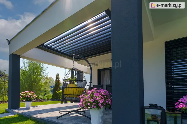 Widok na nowoczesny taras z zamontowanym w opasce betonowej wspartej na szarym masywnym słupie dachem lamelowym SB400R. Aluminiowa rama dachu w kolorze antracytowym jest wbudowana w otwór białego stropu, a jej ruchome pióra (lamele) są ustawione pod kątem, co pozwala na częściowe przepuszczanie światła słonecznego. Pod zadaszeniem znajduje się strefa wypoczynkowa z wiszącym fotelem typu kokon na czarnym stelażu oraz meblami tarasowymi w tle. Posadzka tarasu wyłożona jest dużymi, szarymi płytami betonowymi. Aranżację uzupełniają donice z kwitnącymi, różowymi kwiatami. W tle widoczny jest zadbany ogród z zielonym trawnikiem oraz gęstą roślinnością i drzewami pod pogodnym, błękitnym niebem. Na elewacji budynku po prawej stronie zamontowane są dopasowane kolorystycznie żaluzje fasadowe.