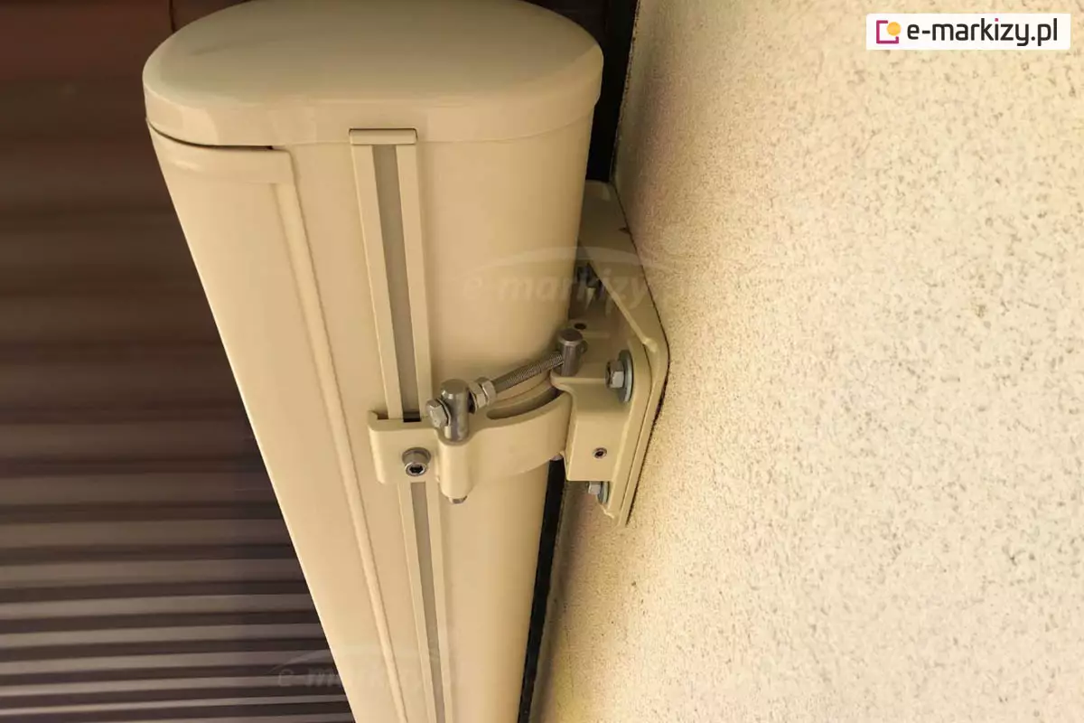 Biała kaseta markizy zamocowana w uchwycie ściennym połączonym z sufitowym. Specjalny rowek w dolnej części kasety zapewnia solidne mocowanie markizy w uchwycie. Jasna markiza zamocowana do kremowej ściany pod sufitem wyłożonym brązowymi panelami.