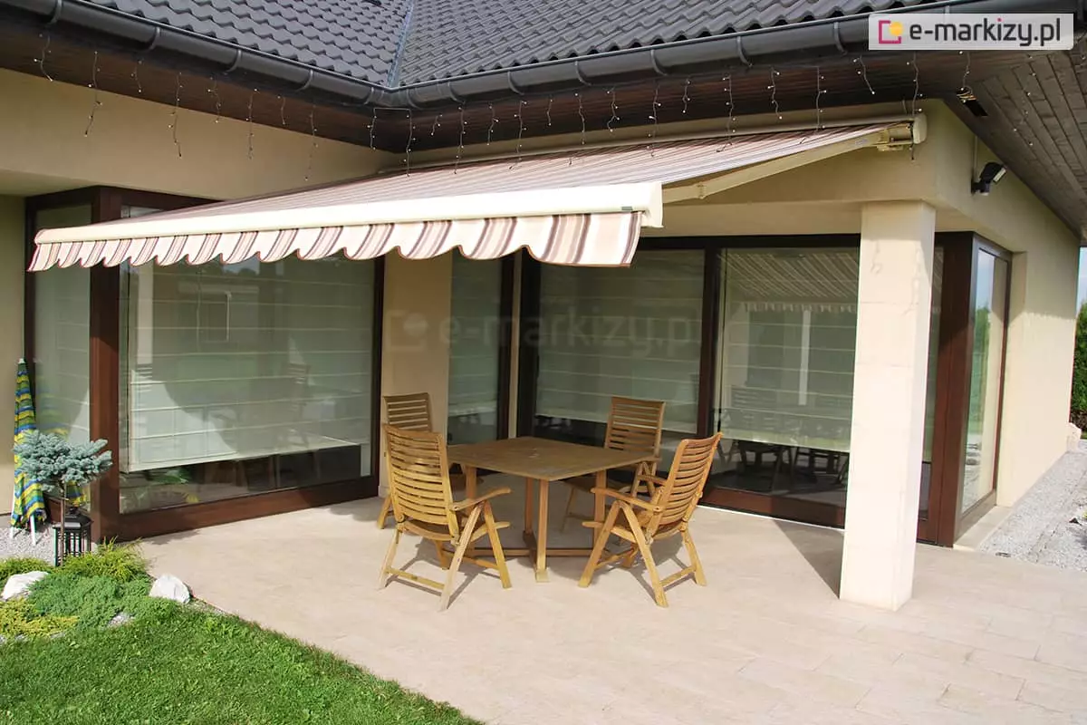 Zdjęcie prezentuje w pełni rozwiniętą markizę tarasową, zamontowaną pod podbitką dachu nowoczesnego domu jednorodzinnego o beżowej elewacji. Poszycie markizy wykonane jest z wysokiej jakości tkaniny w klasyczny wzór pionowych pasów w odcieniach beżu, brązu i bieli, zakończone ozdobną falbaną o falistym brzegu z ciemniejszą lamówką. Konstrukcja markizy opiera się na ramionach przegubowych w kolorze kremowym, które stabilnie utrzymują materiał nad przestrzenią wypoczynkową. Pod zadaszeniem znajduje się zestaw mebli ogrodowych wykonanych z jasnego drewna, składający się z kwadratowego stołu oraz czterech krzeseł z wysokimi oparciami, ustawionych na tarasie wyłożonym jasnymi płytami kamiennymi. W tle widoczne są duże przeszklenia okienne w ciemnobrązowych ramach, w których odbija się otoczenie oraz spód markizy, a wewnątrz pomieszczeń dostrzec można jasne rolety rzymskie. Budynek posiada dach pokryty ciemną dachówką z zamontowanym systemem rynnowym, pod którym zawieszono dekoracyjne sznury lampek zewnętrznych. Po lewej stronie kadru widoczny jest fragment zadbanego ogrodu z zielonym trawnikiem, niskimi nasadzeniami iglaków oraz dekoracyjnym żwirem.
