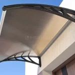 Zdjęcie prezentuje pojedynczy moduł daszka naddrzwiowego o łukowatym kształcie, zamontowany nad wejściem do budynku na tle bezchmurnego, niebieskiego nieba. Konstrukcja wspierająca składa się z dwóch czarnych, ażurowych wsporników wykonanych z wytrzymałego tworzywa polimerowego o wzmocnionej strukturze żeberkowej, które są przytwierdzone do jasnej elewacji o tynkowanej strukturze. Wypełnienie zadaszenia stanowi płyta z poliwęglanu komorowego w odcieniu dymionego brązu, charakteryzująca się widoczną strukturą wewnętrznych kanałów. Krawędzie płyty zabezpieczone są aluminiowymi listwami wykończeniowymi w kolorze czarnym, które usztywniają konstrukcję i chronią brzegi przed czynnikami zewnętrznymi. Pod daszkiem widoczny jest fragment wnęki drzwiowej z elementami wykończonymi materiałem drewnopodobnym oraz fragment ciemnego skrzydła drzwiowego. Perspektywa zdjęcia ujęta jest od dołu, co podkreśla krzywiznę profilu i detale montażowe wsporników.