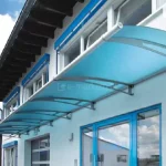 Nowoczesny system zadaszenia Elegant wykonany z akrylu satynowanego w kolorze niebieskim, zamontowany na elewacji budynku usługowego lub przemysłowego. Konstrukcja składa się z kilku połączonych modułów o łukowym kształcie, tworzących długą, jednolitą osłonę nad niebieskimi, przeszklonymi drzwiami wejściowymi i witrynami. Płyty akrylowe są osadzone na wytrzymałych stalowych wspornikach w kolorze jasnoszarym, które charakteryzują się nowoczesnym designem z geometrycznymi otworami. Budynek posiada białą elewację o tynku strukturalnym, a dach wykończony jest ciemną podbitką i rynnami. W tle, pod intensywnie błękitnym niebem z białymi chmurami typu cumulus, widoczne są fragmenty sąsiedniej zabudowy miejskiej oraz drzewa liściaste. Perspektywa zdjęcia podkreśla lekkość konstrukcji oraz możliwość jej nieograniczonego przedłużania poprzez dodawanie kolejnych modułów.