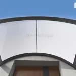 Grafika przedstawia widok z góry na nowoczesny daszek naddrzwiowy o łukowatym kształcie, zamontowany nad wnęką wejściową budynku. Konstrukcja zadaszenia składa się z ciemnoantracytowej ramy aluminiowej, która dzieli powierzchnię na dwa symetryczne panele. Wypełnienie paneli stanowi biały, półtransparentny poliwęglan komorowy, który skutecznie rozprasza światło. Rama daszka posiada wyraźnie zaznaczone profile krawędziowe oraz pionowy szpros przebiegający dokładnie przez środek konstrukcji. Daszek jest zamontowany do jasnej, płaskiej elewacji i wystaje poza obrys wnęki drzwiowej, widocznej w dolnej części kadru. Wnęka ta ukazuje górny fragment drzwi o brązowym kolorze z pionowym, ciemniejszym pasem dekoracyjnym. Całość prezentowana jest na tle czystego, błękitnego nieba z delikatnymi smugami białych chmur po bokach. Perspektywa od dołu pozwala dokładnie zaobserwować łukowate wygięcie daszka oraz sposób, w jaki jego krawędzie są wykończone profilami odprowadzającymi wodę opadową na boki.