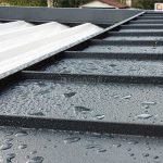 Zdjęcie przedstawia szczegółowe zbliżenie na górną część konstrukcji pergoli Luxury po opadach deszczu, koncentrując się na systemie ochrony poszycia. Na pierwszym planie widoczny jest stały daszek ochronny wykonany z profilowanej blachy w kolorze ciemnografitowym o matowej, ziarnistej strukturze, na której powierzchni zalegają liczne krople wody. Daszek ten składa się z równoległych koryt odprowadzających wodę, zamontowanych prostopadle do ramy pergoli. Po lewej stronie widoczny jest fragment jasnego, białego poszycia materiałowego dachu, które jest częściowo schowane pod metalową osłoną. Materiał zadaszenia ma delikatnie falistą strukturę i również jest pokryty wilgocią. W tle, poza krawędzią konstrukcji, widać rozmyty zarys ogrodu oraz dachy sąsiednich budynków pod zachmurzonym niebem. Ujęcie to dokumentuje skuteczność odprowadzania wody z systemowych elementów osłonowych pergoli.