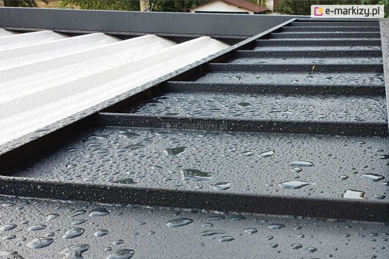 Zdjęcie przedstawia szczegółowe zbliżenie na górną część konstrukcji pergoli Luxury po opadach deszczu, koncentrując się na systemie ochrony poszycia. Na pierwszym planie widoczny jest stały daszek ochronny wykonany z profilowanej blachy w kolorze ciemnografitowym o matowej, ziarnistej strukturze, na której powierzchni zalegają liczne krople wody. Daszek ten składa się z równoległych koryt odprowadzających wodę, zamontowanych prostopadle do ramy pergoli. Po lewej stronie widoczny jest fragment jasnego, białego poszycia materiałowego dachu, które jest częściowo schowane pod metalową osłoną. Materiał zadaszenia ma delikatnie falistą strukturę i również jest pokryty wilgocią. W tle, poza krawędzią konstrukcji, widać rozmyty zarys ogrodu oraz dachy sąsiednich budynków pod zachmurzonym niebem. Ujęcie to dokumentuje skuteczność odprowadzania wody z systemowych elementów osłonowych pergoli.