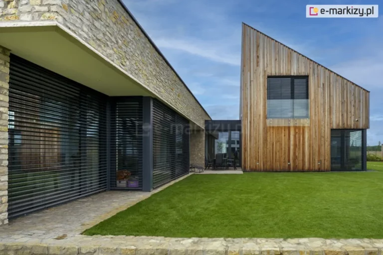 Designerski dom jednorodzinny składający się z dwóch połączonych części - parterowej z fasadą z kamienia oraz dużymi oknami oraz piętrowej o skośnym dachu. którego elewację wyłożono drewnem. Wszystkie okna budynków osłonięte zostały żaluzjami fasadowymi podtynkowymi Z90 Box w kolorze antracytowym, który współgra z kolorami wykończenia okien. Przed budynkiem widoczny zadbany trawnik oraz ścieżki wyłożone kamieniami. Nas wszystkim widoczne niebieskie, lekko zachmurzone niebo.