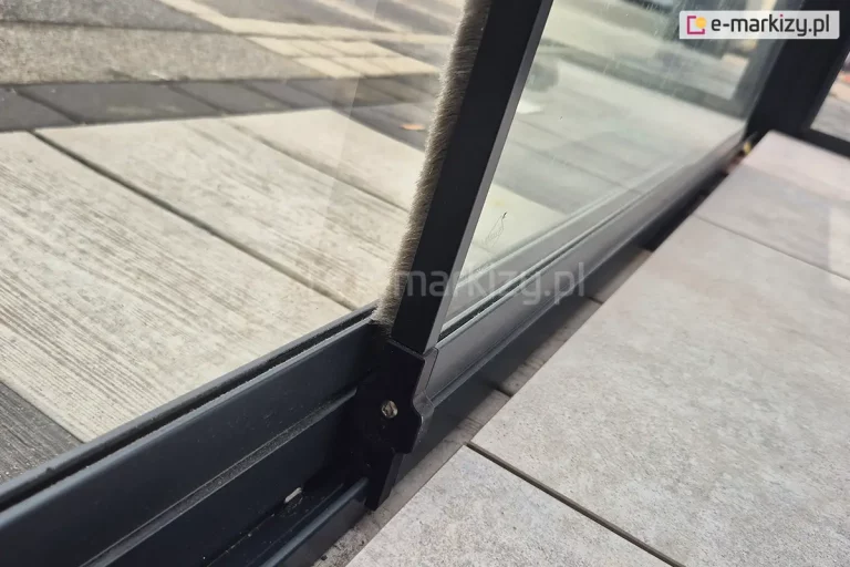 Zbliżenie boku skrzydła przesuwnego systemu szklanego Miros. Widoczny panel dolny osadzony na prowadnicy oraz listwa boczna z uszczelka z włosia. Za panelem widoczna kolejna tafla szkła, która mija się z widocznym panelem. Całość zamocowana na drewnianym tarasie, wyłożonym przed szybami dodatkowymi płytkami.