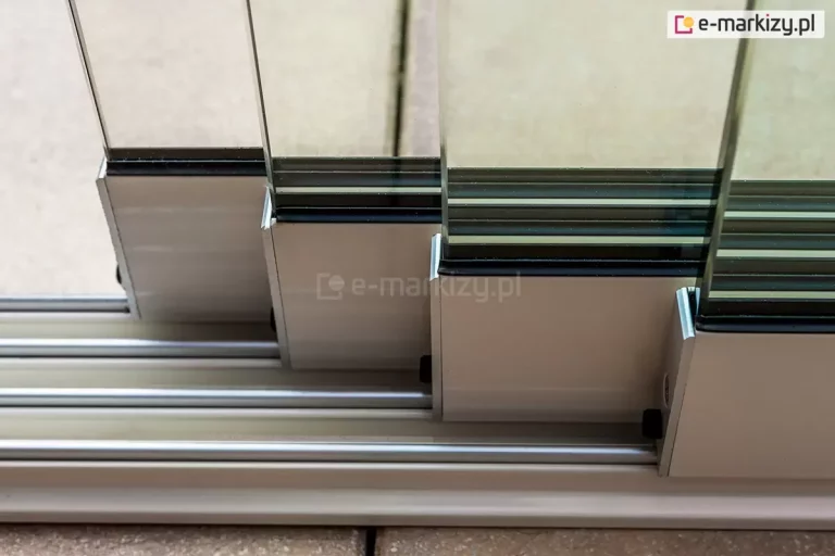 Zbliżenie techniczne na dolny system jezdny szklanej ściany przesuwnej Auris. Zdjęcie przedstawia cztery tafle bezramowego szkła hartowanego osadzone w aluminiowych profilach dolnych w kolorze srebrnym/satynowym. Każdy panel szklany spoczywa na osobnej szynie niskoprogowej prowadnicy wielotorowej. Widoczny jest sposób montażu szkła w profilu za pomocą czarnych uszczelek stabilizujących oraz kaskadowe ułożenie paneli, które umożliwia ich swobodne przesuwanie względem siebie. Prowadnica jest zamontowana bezpośrednio na podłożu tarasowym, zapewniając minimalny próg i płynną pracę wózków jezdnych.
