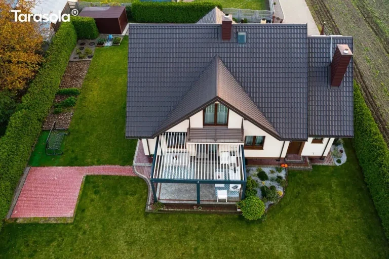 Zdjęcie wykonane prosto z góry (widok typu top-down) przedstawia bryłę nowoczesnego domu jednorodzinnego z zainstalowaną dwumodułową pergolą Essential Tarasola. Budynek posiada wielospadowy dach pokryty ciemnobrązową dachówką, z widocznymi kominami oraz oknami dachowymi. Aluminiowa konstrukcja Essential Tarasola w kolorze antracytowym przylega do jednej z elewacji, a jej dach tworzą białe, równolegle ułożone lamele, które w tej perspektywie przypominają jasne pasy. Pod zadaszeniem widoczna jest aranżacja tarasu z podziałem na część jadalnianą oraz wypoczynkową, ułożona na jasnej kostce brukowej. Z tej wysokości wyraźnie widać zagospodarowanie terenu wokół domu. Od tarasu w lewą stronę prowadzi ścieżka wykonana z czerwonej kostki brukowej, kończąca się placem, na którym ustawiono huśtawkę ogrodową. Całość posesji otoczona jest gęstym, zielonym żywopłotem, który wyznacza granice działki i oddziela ją od sąsiednich terenów uprawnych oraz drogi dojazdowej. Ogród składa się z dużej połaci zadbanego trawnika oraz dekoracyjnej rabaty roślinnej wysypanej jasnym kruszywem, przylegającej bezpośrednio do narożnika tarasu z pergolą Essential Tarasola. W górnej części kadru widoczny jest mniejszy budynek gospodarczy oraz uporządkowana strefa z grządkami uprawnymi. Zdjęcie doskonale obrazuje, jak system Essential Tarasola integruje się z architekturą domu i planem całego ogrodu.