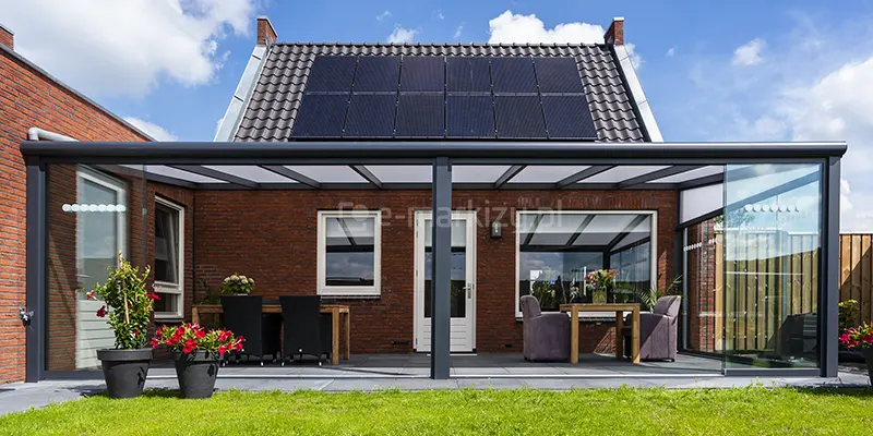 Frontowy widok na nowoczesne, szerokie zadaszenie tarasowe Lunaro z wypełnieniem z poliwęglanu, zamontowane przy parterowym budynku z czerwonej cegły. Konstrukcja w kolorze antracytowym posiada jednospadowy dach z mlecznymi panelami poliwęglanowymi oraz częściową zabudowę szklaną (dwie części frontu oraz prawy bok), na zsuniętych taflach ze szkła hartowanego widoczne poziome pasy białych, okrągłych emblematów. Pod zadaszeniem, na tarasie wyłożonym szarymi płytami, zaaranżowano dwie strefy wypoczynkowe: po lewej stronie znajduje się drewniany stół z czarnymi krzesłami, a po prawej dwa fioletowe, tapicerowane fotele przy mniejszym stoliku. Na pierwszym planie, przy krawędzi zielonego trawnika, ustawiono dwie duże czarne donice z kwitnącymi czerwonymi kwiatami. Budynek posiada białe ramy okienne i drzwiowe, a na jego spadzistym dachu z ciemnej dachówki zainstalowano duży system paneli fotowoltaicznych.