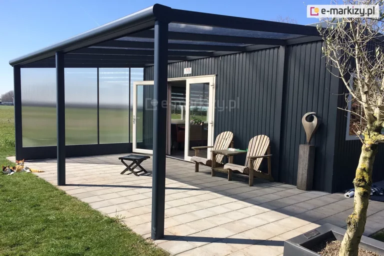 Nowoczesne, przyścienne zadaszenie tarasowe zamontowane przy elewacji domku holenderskiego wykończonego ciemną, pionową blachą trapezową. Konstrukcja zadaszenia wykonana jest z profili aluminiowych w kolorze antracytowym. Dach posiada lekki spadek i jest pokryty przezroczystymi płytami z poliwęglanu, które są osadzone w systemowych profilach dachowych. Całość wspiera się na solidnych, okrągłych słupach nośnych umieszczonych na krawędzi tarasu wykonanego z jasnych, kwadratowych płyt betonowych. Lewa strona zadaszenia została zabudowana boczną ścianką z poliwęglanu komorowego o mlecznym, matowym wykończeniu, co zapewnia prywatność i osłonę przed wiatrem. Pod zadaszeniem znajdują się dwa drewniane krzesła ogrodowe ze stolikiem kawowym, ustawione obok szerokich, białych drzwi tarasowych prowadzących do wnętrza domku. W tle widoczny jest otwarty teren zielony oraz błękitne, bezchmurne niebo.