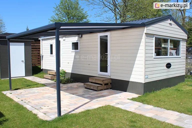 Zdjęcie ukazuje jasny domek holenderski o elewacji z poziomych paneli, do którego zamontowano przyścienne zadaszenie tarasowe Nebbiolo marki Deponti. Konstrukcja zadaszenia opiera się na ciemnoszarym, aluminiowym stelażu, który przylega bezpośrednio do bocznej ściany domku pod linią dachu i jest wsparty z przodu na dwóch pionowych słupach o kwadratowym przekroju. Dach wiaty wykonany jest z mlecznych płyt poliwęglanowych, które rzucają lekki cień na przestrzeń wejściową. Teren pod zadaszeniem oraz ścieżka prowadząca do domku zostały utwardzone kostką brukową w jasnych, piaskowych kolorach. Przy wejściach do domku znajdują się niskie, drewniane schodki. W tle widoczny jest mniejszy budynek gospodarczy z białymi drzwiami, zielony trawnik oraz wysokie drzewa liściaste pod błękitnym niebem.