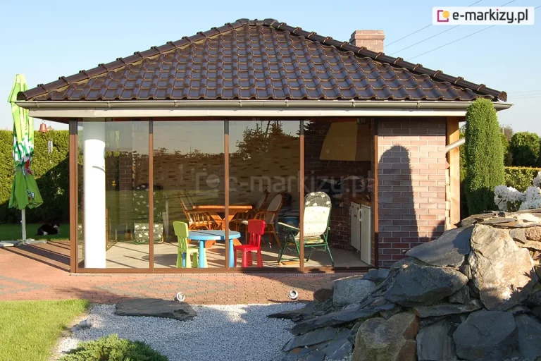 Murowany domek ogrodowy z dachem z ciemnej dachówki, którego front został zabudowany systemem szklanym Sotro. Ściana frontowa składa się z czterech wysokich paneli szklanych w cienkich ramach o kolorze dopasowanym do ceglanej elewacji. Wewnątrz widać strefę jadalnianą z drewnianym stołem, krzesłami oraz kącikiem dla dzieci z kolorowymi meblami. Zabudowa znajduje się na podłożu z kostki brukowej, a przed nią umieszczono punktowe reflektory oświetleniowe skierowane na konstrukcję. Po prawej stronie na pierwszym planie widoczny wysoki skalniak ułożony z dużych głazów. Po lewej stronie zadaszenia widoczny złożony parasol ogrodowy