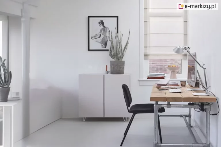 Zdjęcie przedstawia jasne, nowocześnie urządzone domowe biuro w minimalistycznym stylu. Głównym elementem dekoracyjnym okna jest roleta rzymska wykonana z półtransparentnego, kremowego materiału z widocznymi poziomymi fiszbinami, które nadają jej regularną strukturę. Roleta jest częściowo podciągnięta, odsłaniając dolną część okna, przez które widać ceglaną elewację sąsiedniego budynku. Na parapecie pod oknem leży stos złożonych tkanin oraz stoi dekoracyjna misa na wysokiej nóżce. Po prawej stronie znajduje się biurko o industrialnym charakterze, wykonane z grubego, jasnego blatu drewnianego o widocznej strukturze desek, wsparte na stelażu z metalowych rur. Na biurku ustawiony jest monitor komputera, biała klawiatura, srebrna lampka kreślarska oraz drobne akcesoria biurowe. Przy biurku stoi nowoczesne, czarne krzesło na cienkich nogach. Ściany pomieszczenia są białe, a na centralnej ścianie wisi czarno-biała grafika w czarnej ramie przedstawiająca sylwetkę kobiety. Poniżej obrazu znajduje się niska, biała szafka o prostej formie, na której ustawiono donicę z wysokim sukulentem oraz małą świecę. Podłoga w pomieszczeniu jest jasna i jednolita, co podkreśla sterylny i uporządkowany charakter wnętrza. Całość oświetlona jest miękkim, naturalnym światłem wpadającym przez okno.